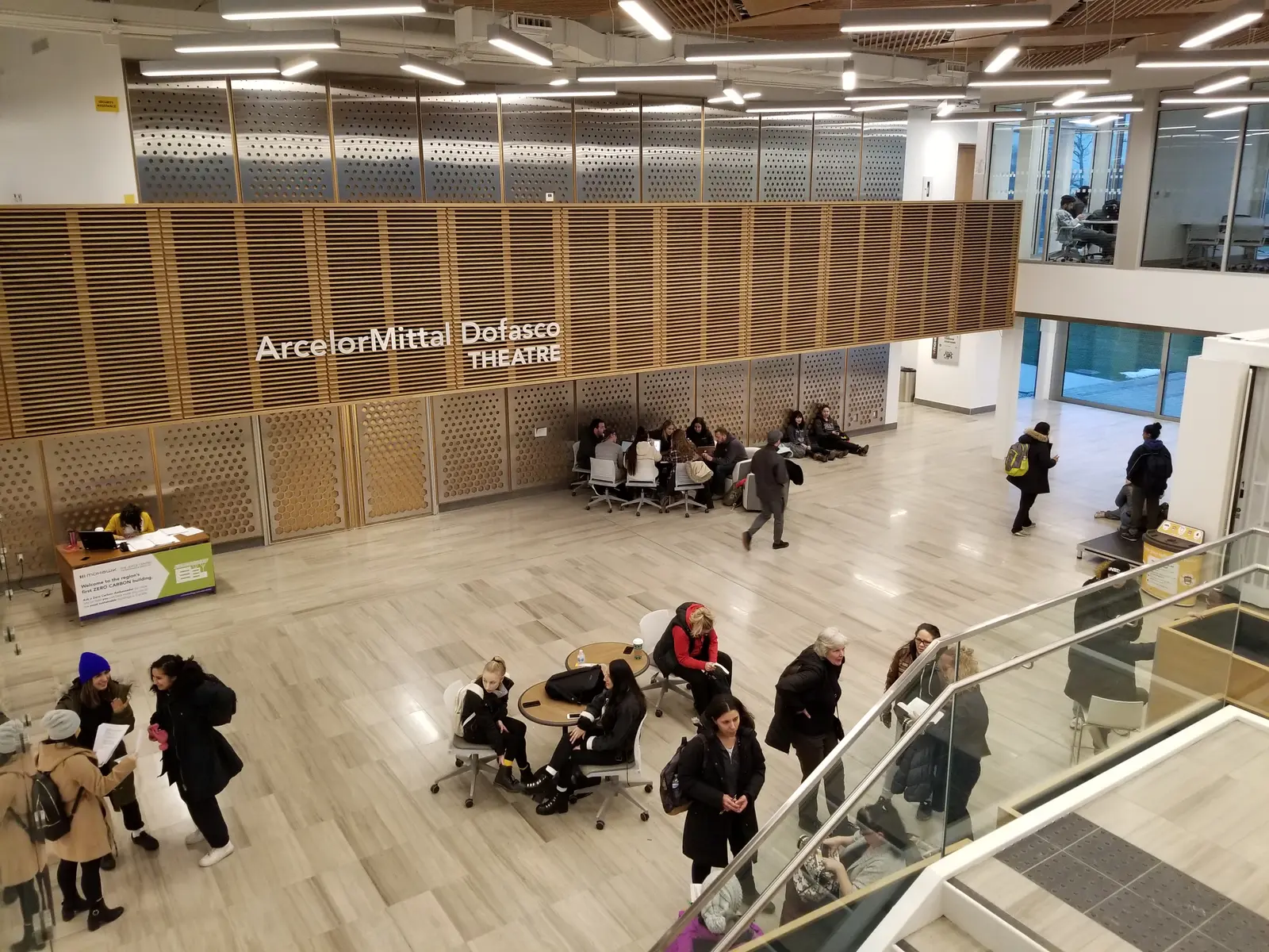 Students sitting and walking in a bright two-storey atrium with wood slat walls and a sign for the ArcelorMittal Dofasco Theatre