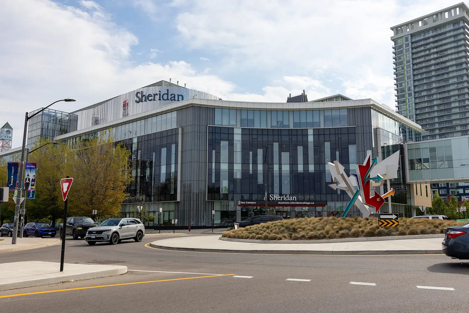 Glass front of Sheridan's Hazel McCallion Campus in Mississauga, with a Sheridan sign and a sculpture on a roundabout