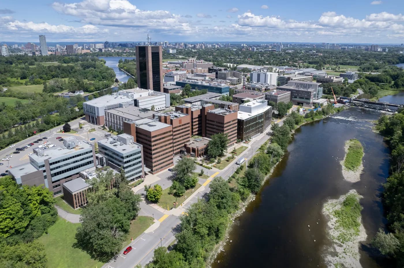 Aerial view of Carleton's campus buildings beside the Rideau River, with a tall dark tower, green space and the Ottawa skyline in the distance