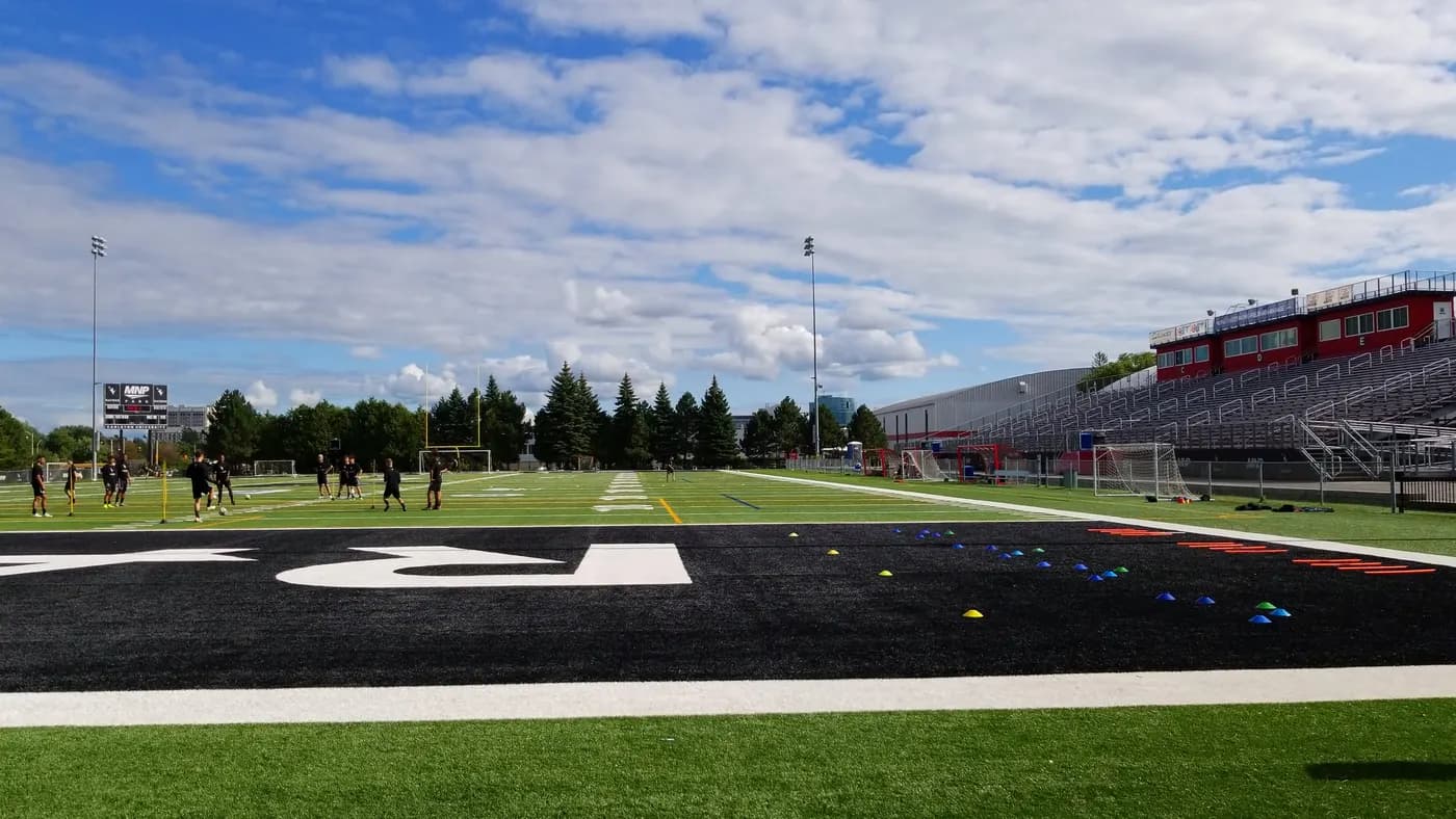 Players warming up on the artificial turf of Carleton's football stadium, with a black end zone, grandstands and a red press box