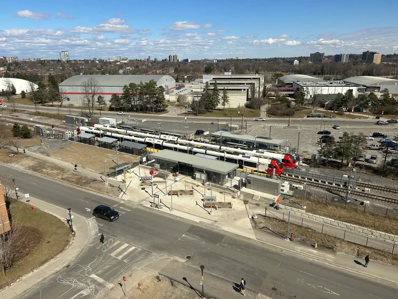Overhead view of Carleton O-Train station with two red-and-white trains at the platforms, a parking lot and campus buildings behind