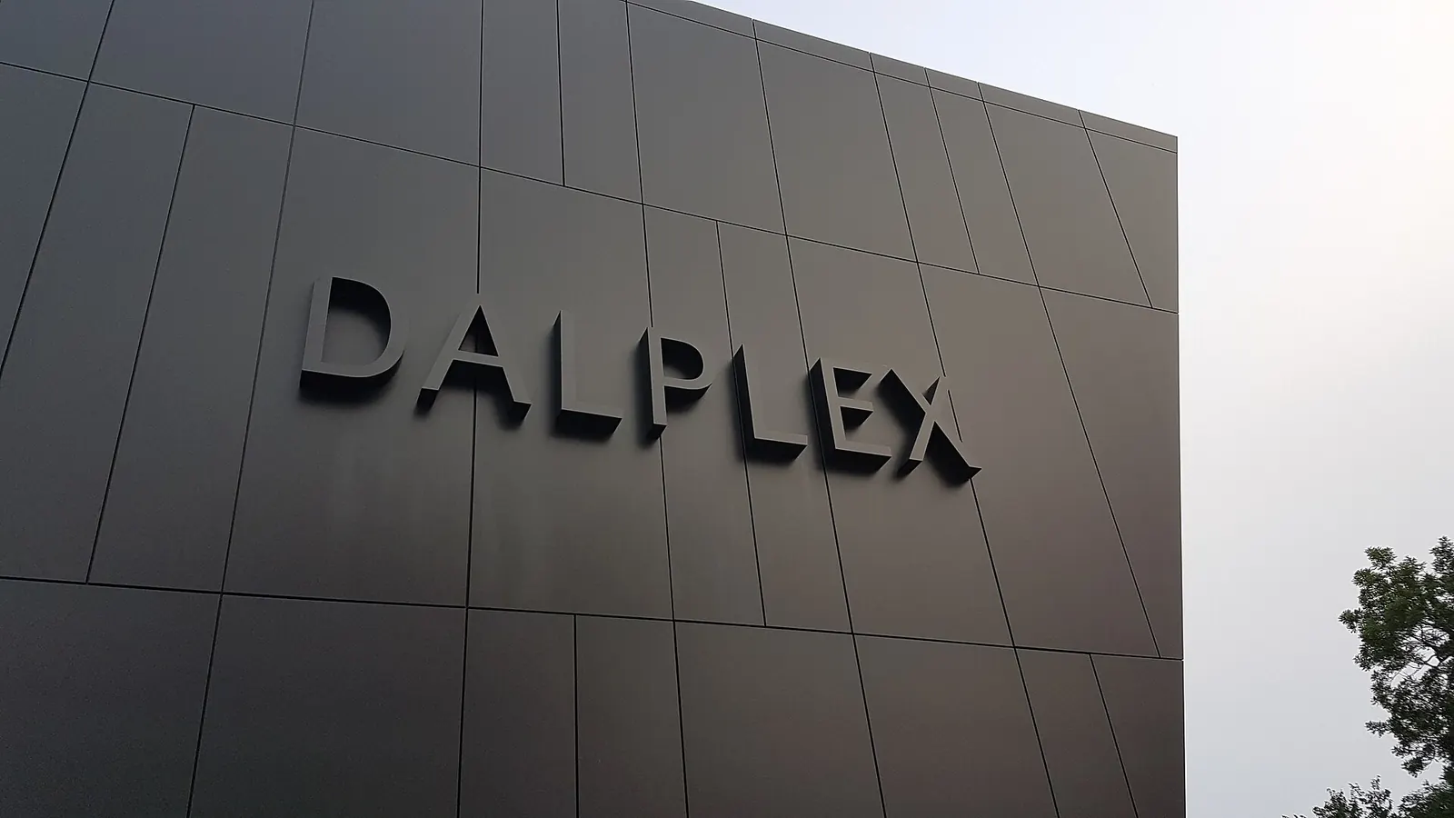 Large raised DALPLEX letters on the dark grey panelled wall of Dalhousie's athletic centre, with a tree at the edge of the frame