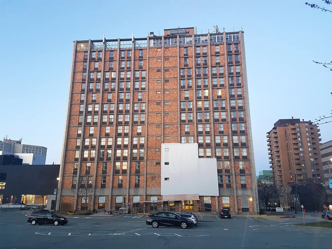 Twelve-storey red-brick Gerard Hall on Dalhousie's Sexton Campus at dusk, with a white-clad entrance block and cars in the parking lot in front