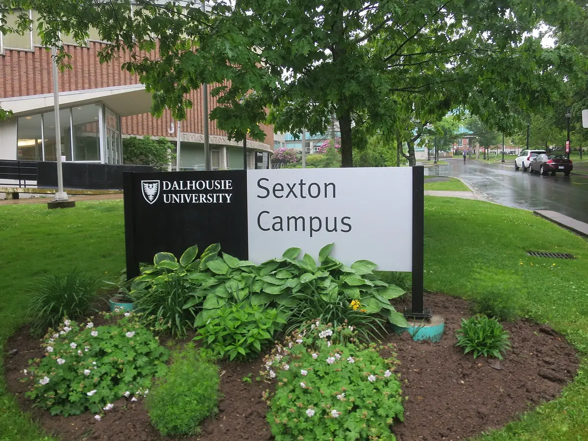 Black and white Dalhousie University Sexton Campus sign in a flower bed, with trees, a wet street and a brick building behind