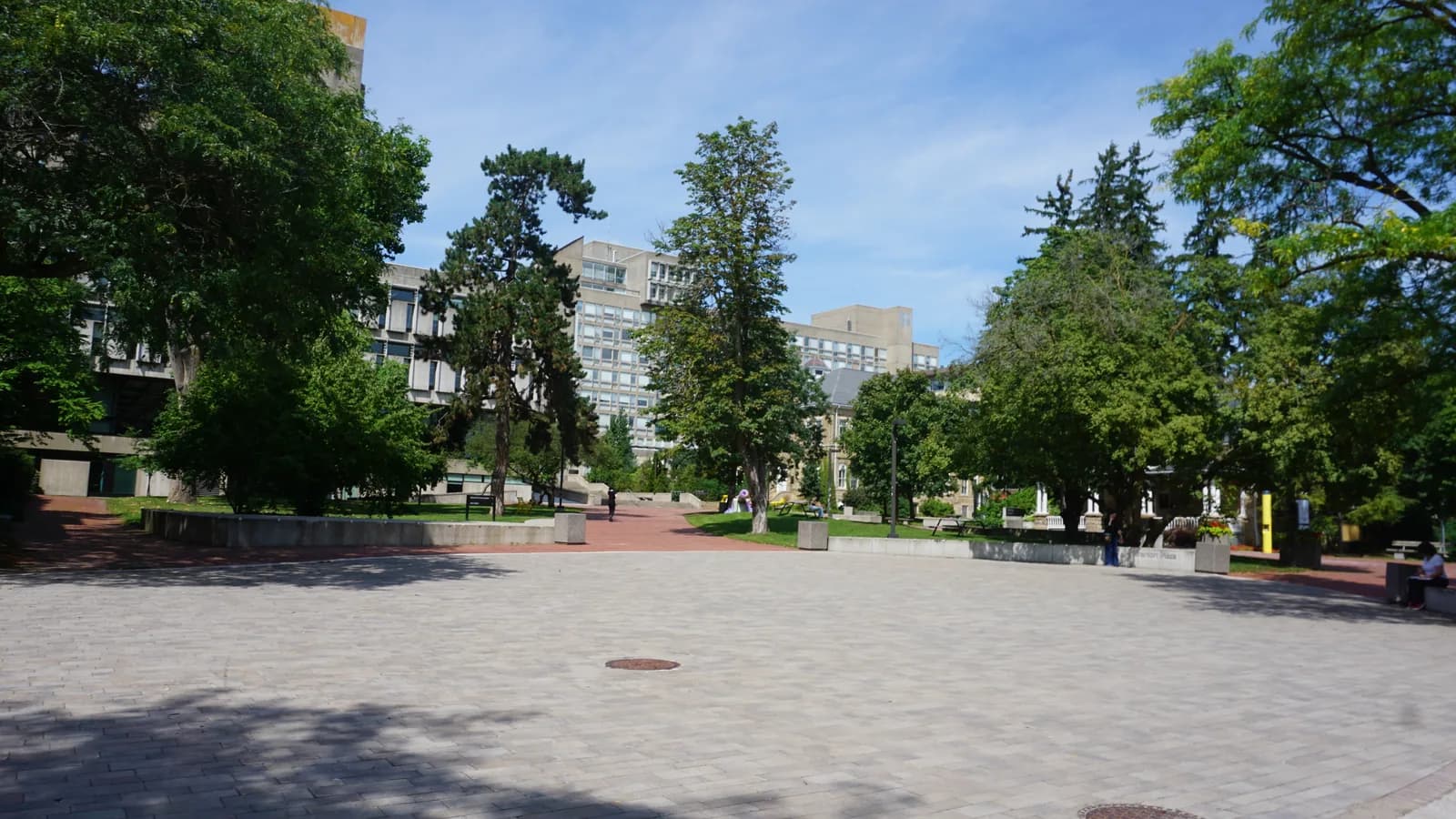 Open paved Branion Plaza on Guelph's main campus with low concrete planters, mature trees and multi-storey concrete buildings behind