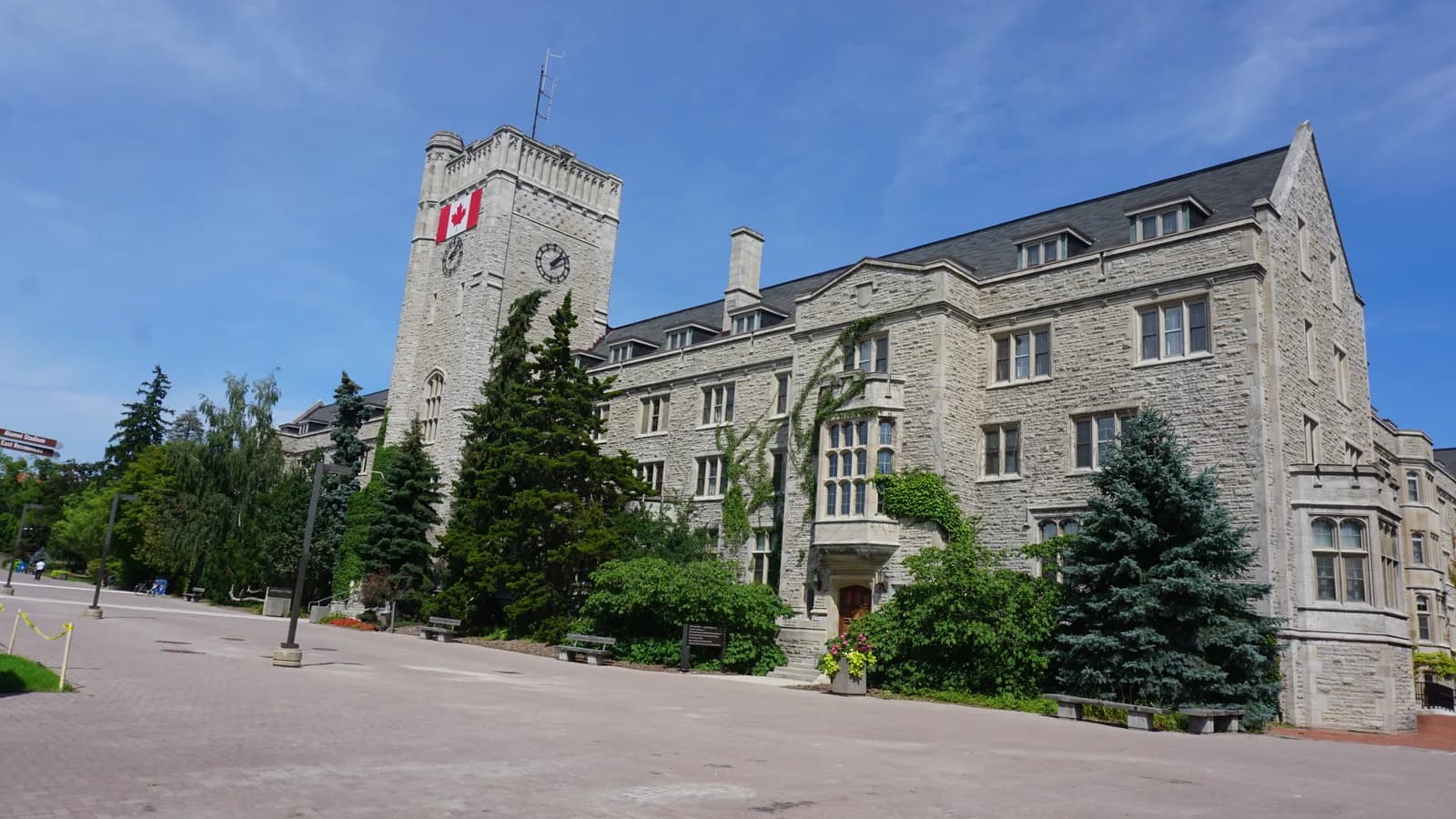 Grey stone Johnston Hall at Guelph with a square clock tower flying a Canadian flag, ivy on the walls and a wide brick walkway in front