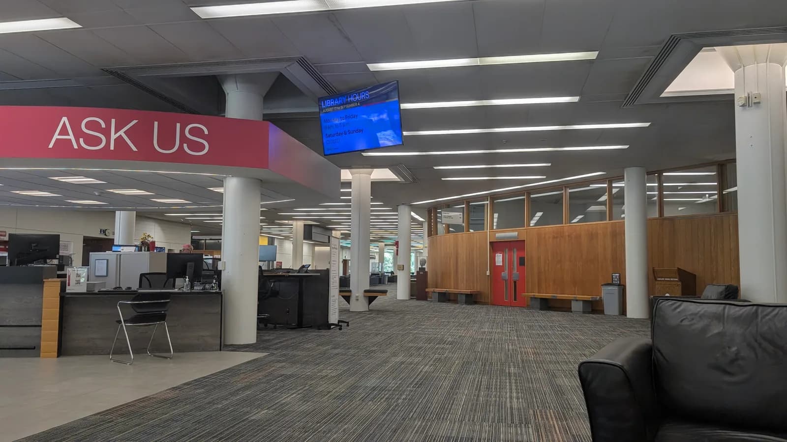 Open floor inside McLaughlin Library with a red Ask Us service desk, white columns, wood-panelled walls and a screen showing library hours
