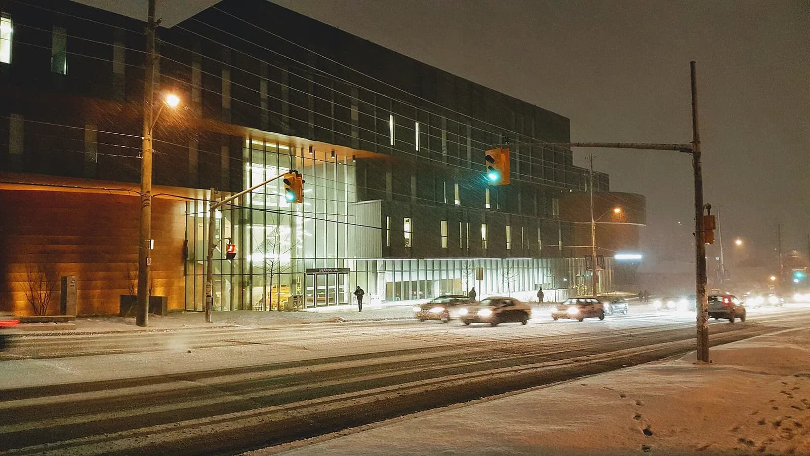 Lazaridis Hall on Laurier's Waterloo campus lit up at night in falling snow, with cars on the street in front