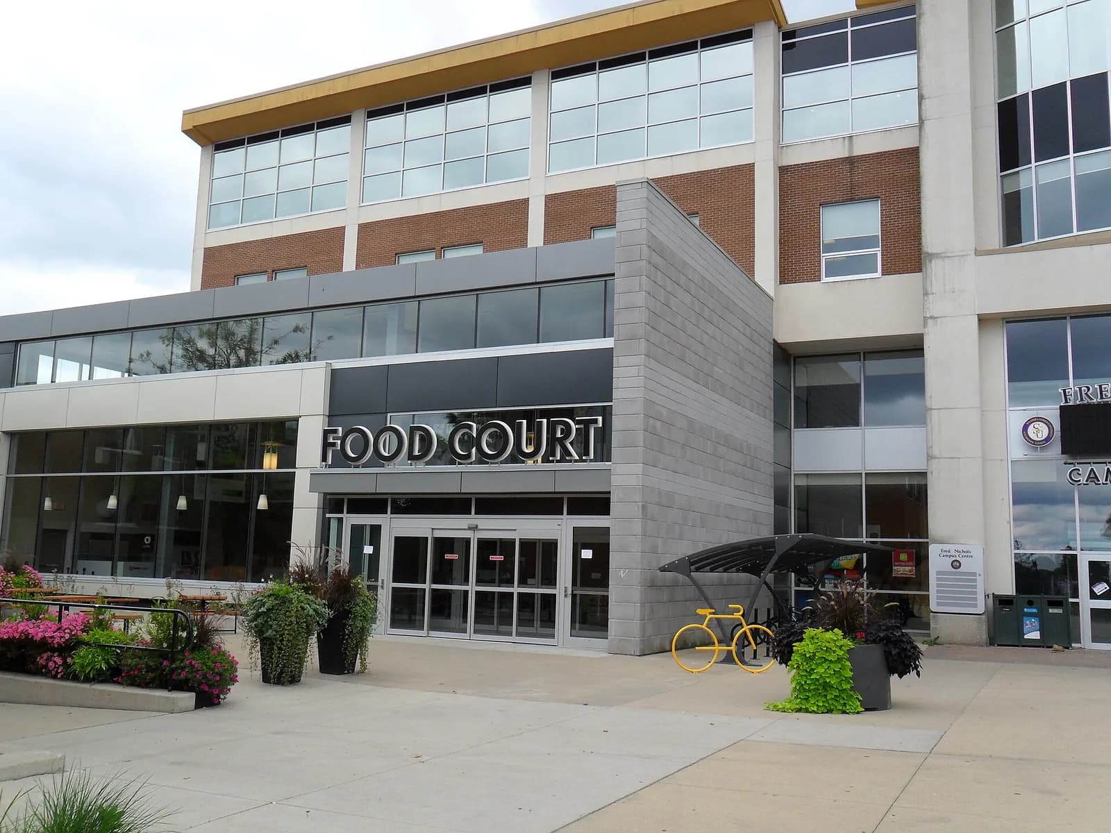 Glass entrance with a Food Court sign on Laurier's Waterloo campus, with planters and a bike shelter outside
