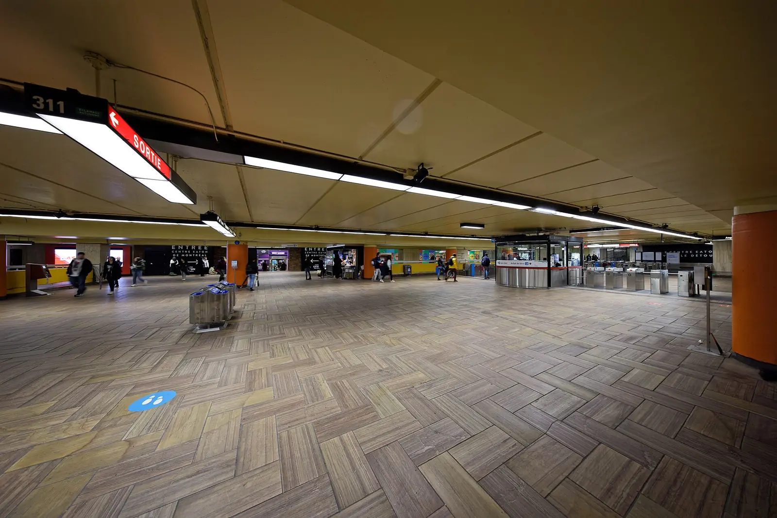 Wide underground concourse of McGill metro station with fare gates, a customer service booth and exit signs
