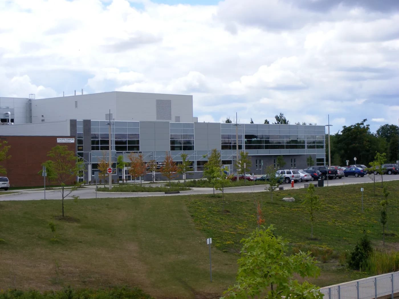 Two-storey grey and glass Campus Recreation and Wellness Centre with an Athletic Complex sign, a parking lot and a grassy slope in front