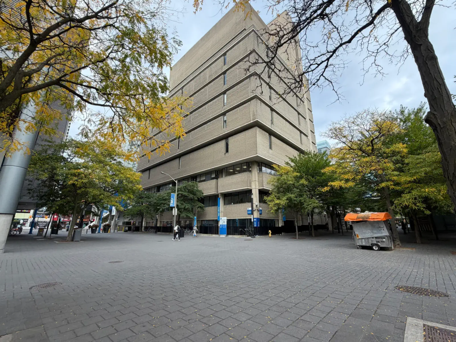 Tall concrete TMU Library Building above a brick-paved plaza with autumn trees and blue TMU banners