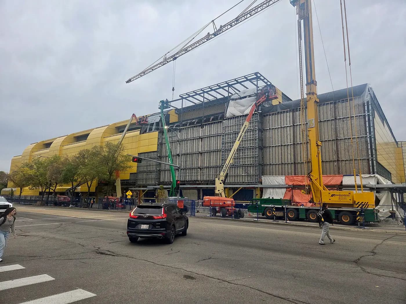The yellow Universiade Pavilion at the University of Alberta wrapped in scaffolding, with a tower crane, boom lifts and construction fencing along the street