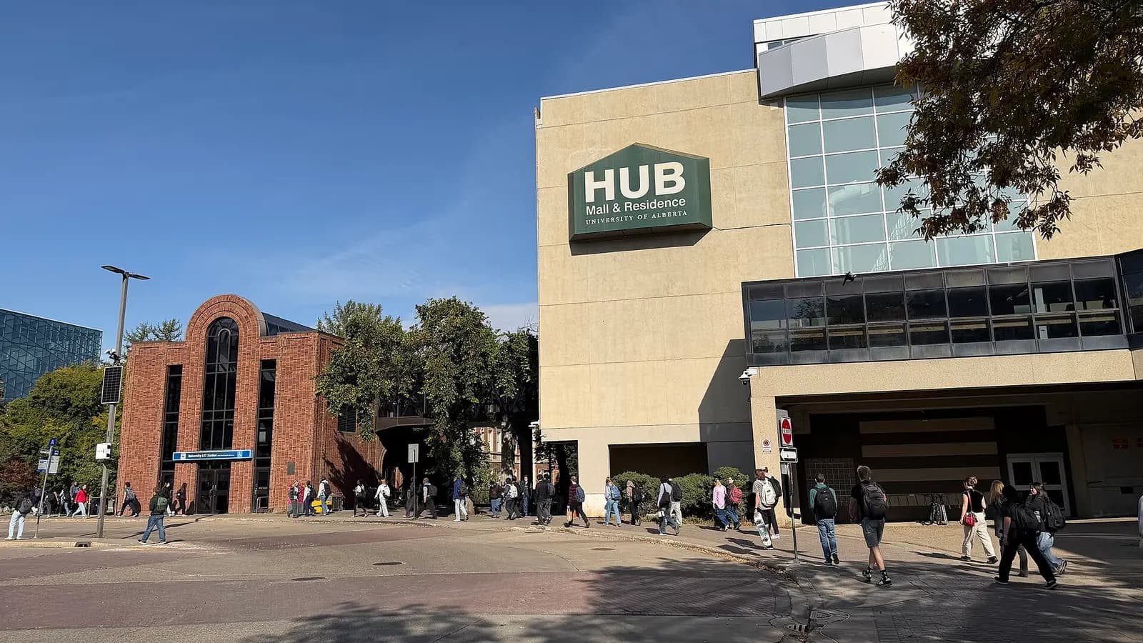 Students walking past the red-brick University LRT station entrance and the HUB Mall and Residence building on North Campus on a sunny day