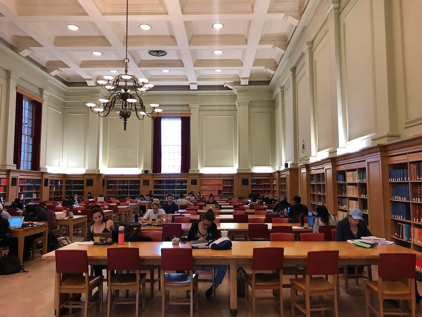 Students studying at long wooden tables with red chairs in Rutherford South's high-ceilinged reading room, with a chandelier, tall windows and bookshelves