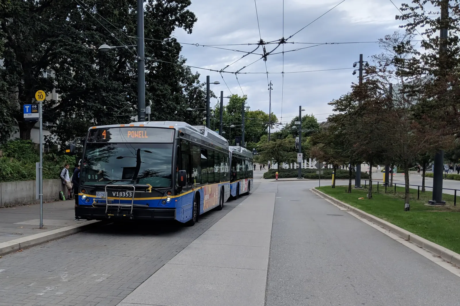 Two TransLink trolley buses waiting at a bay in the UBC Exchange bus loop, with students on the sidewalk