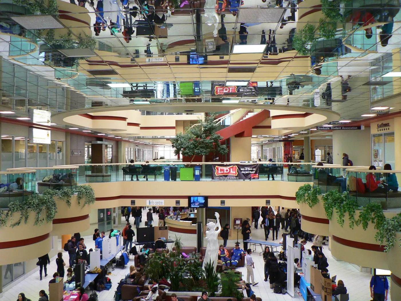 Busy three-storey atrium of UCalgary's MacEwan Student Centre with a mirrored ceiling, balconies, an info centre desk and a Last Defence Lounge banner