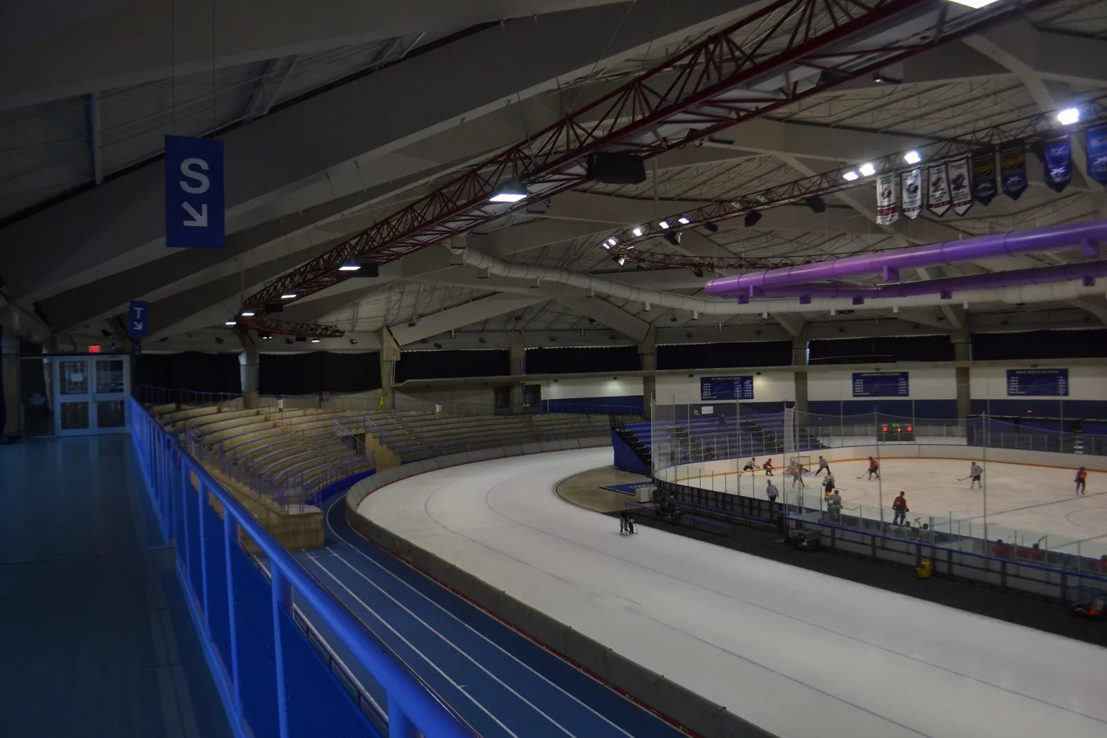 Inside UCalgary's Olympic Oval, with a blue running track and seating on the left, the long-track speed skating ice, and a hockey game on an inner rink