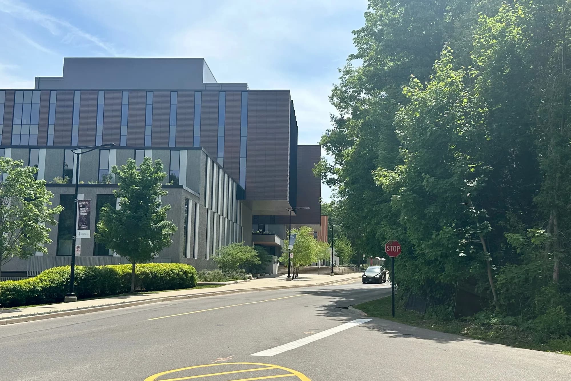 Maanjiwe nendamowinan, a multi-storey brown-panelled academic building beside a tree-lined road at U of T Mississauga