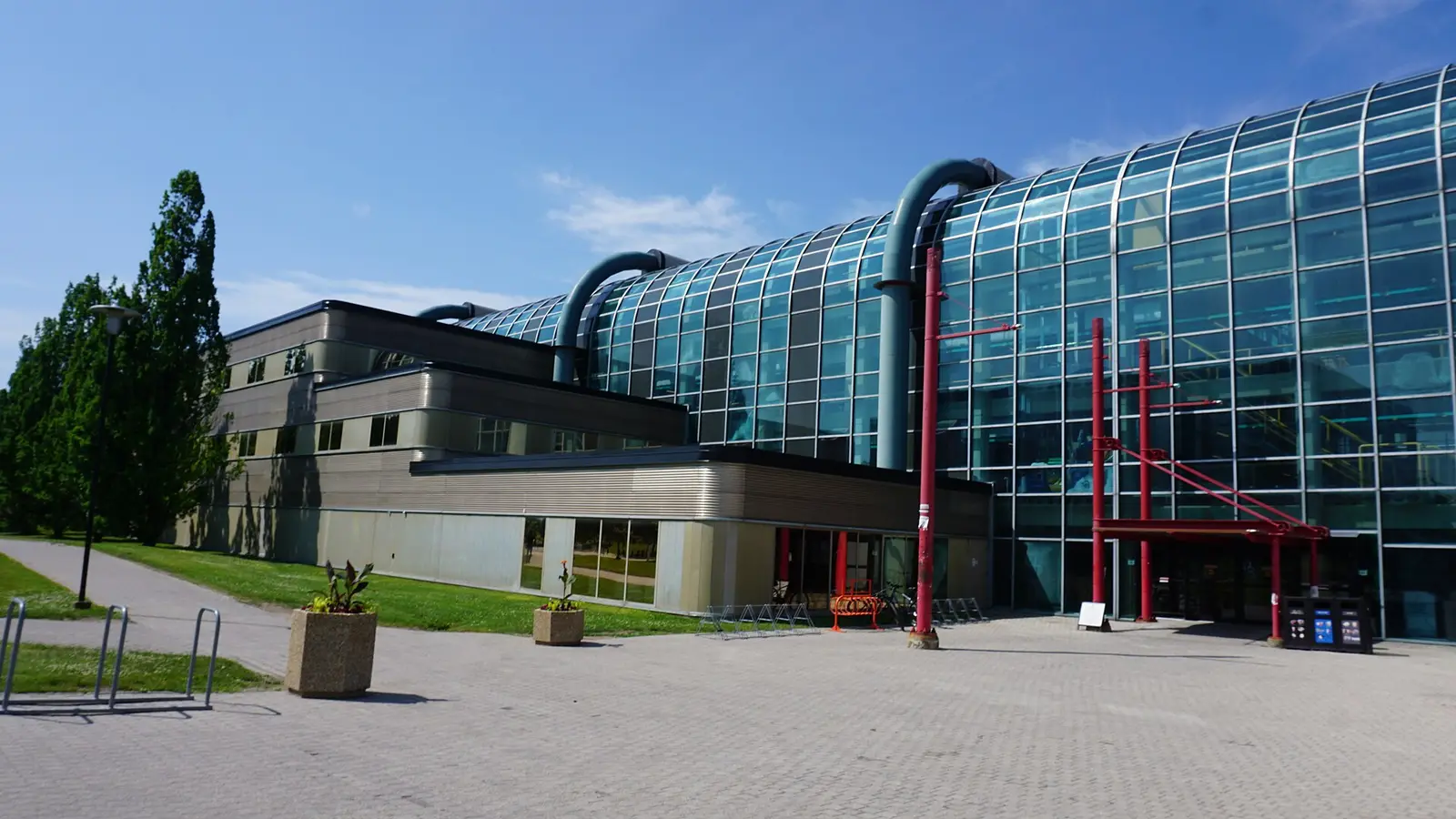 Curved glass and steel exterior of the Davis Centre at the University of Waterloo, with red entrance supports and a paved plaza