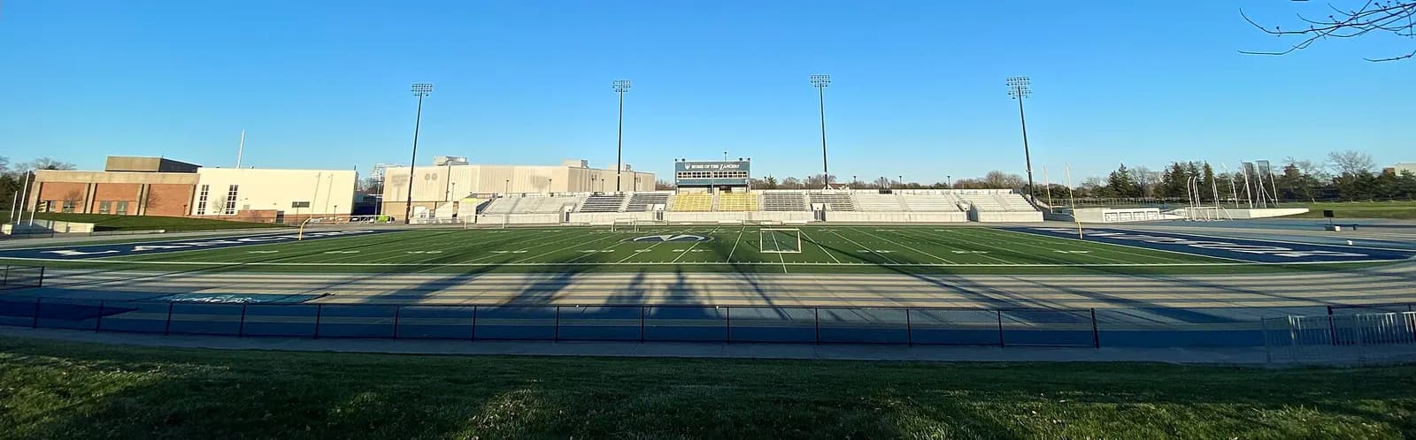 Wide view of UWindsor's Alumni Stadium with a turf football field, a blue running track, grandstands and light towers, and campus buildings behind