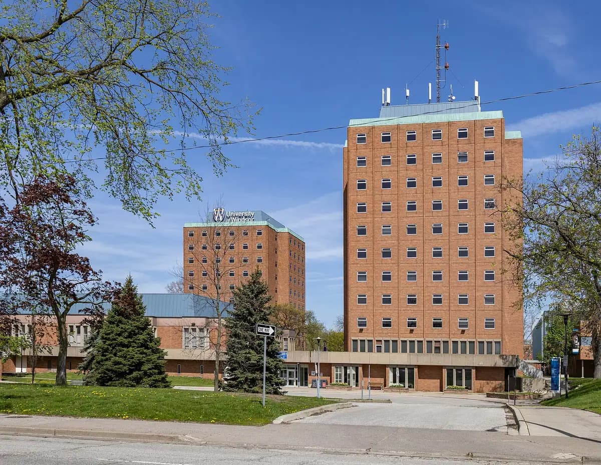 Two tall brick residence towers with green copper trim rising behind a low brick building and lawn at the University of Windsor