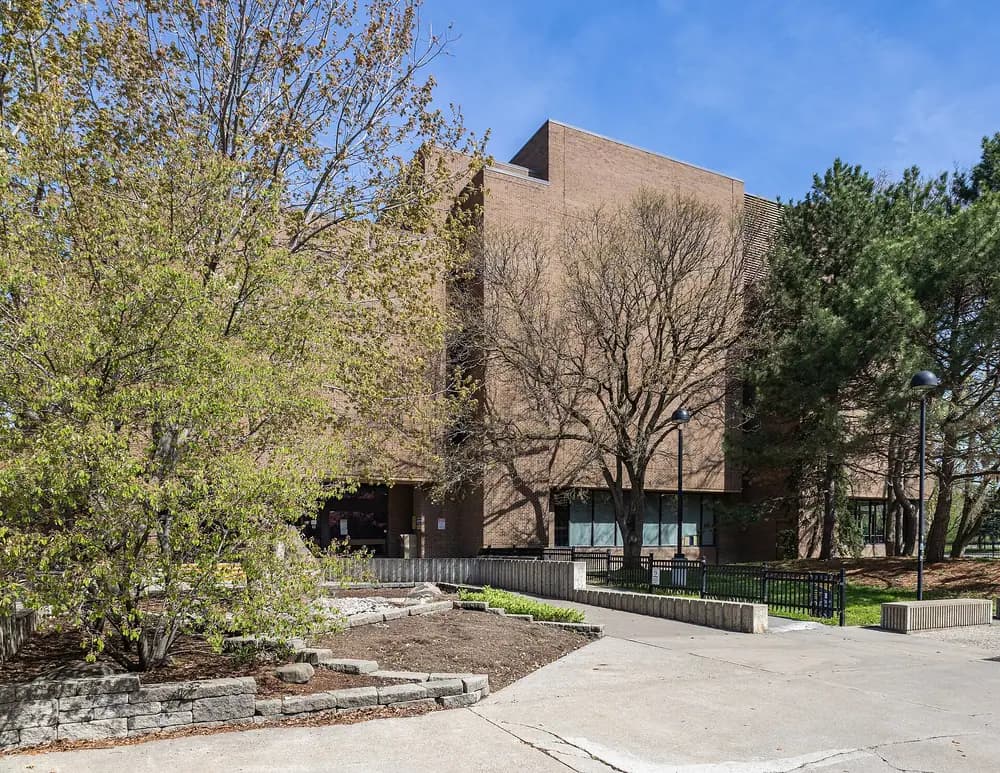 Brown brick exterior of UWindsor's Leddy Library behind spring trees, stone planters and a paved walkway
